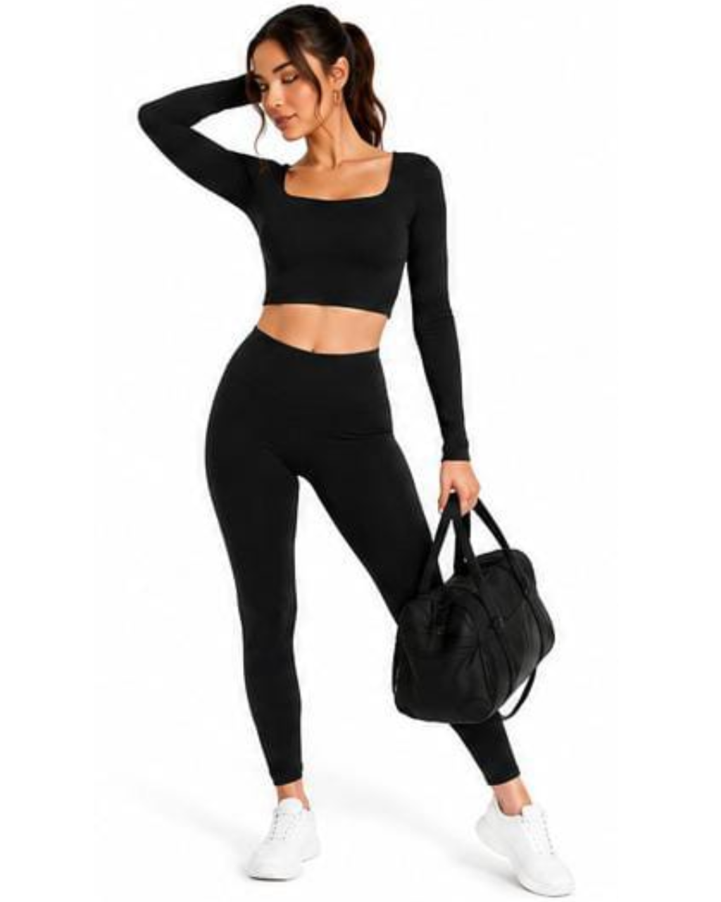 A woman modeling a black activewear set consisting of a long-sleeve crop top and high-waisted leggings, paired with white sneakers and holding a black duffel bag against a plain white background.