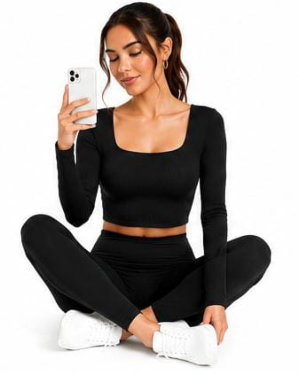 A woman sitting cross-legged on a white background, wearing a black ribbed long-sleeve crop top and matching black leggings, holding a white smartphone.