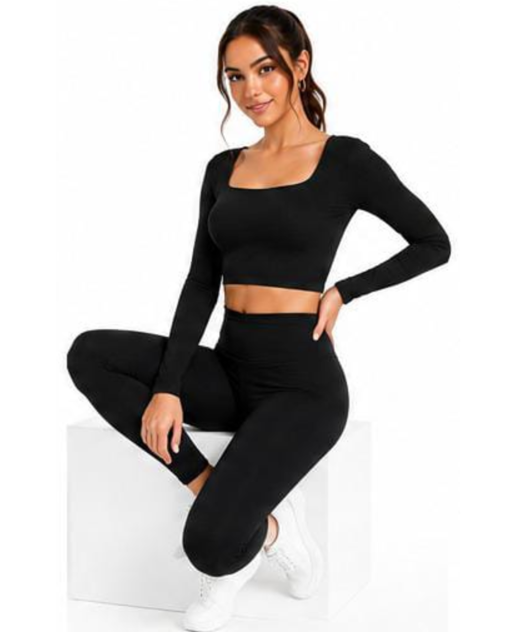 A woman models a matching black activewear set consisting of a long-sleeve crop top and high-waisted leggings, sitting on a white block against a plain white background.