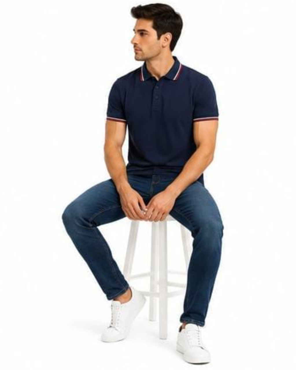 A man wearing a navy blue short-sleeve polo shirt with red and white striped collar and sleeve trim, paired with dark blue jeans and white sneakers, sitting on a white stool against a white background.