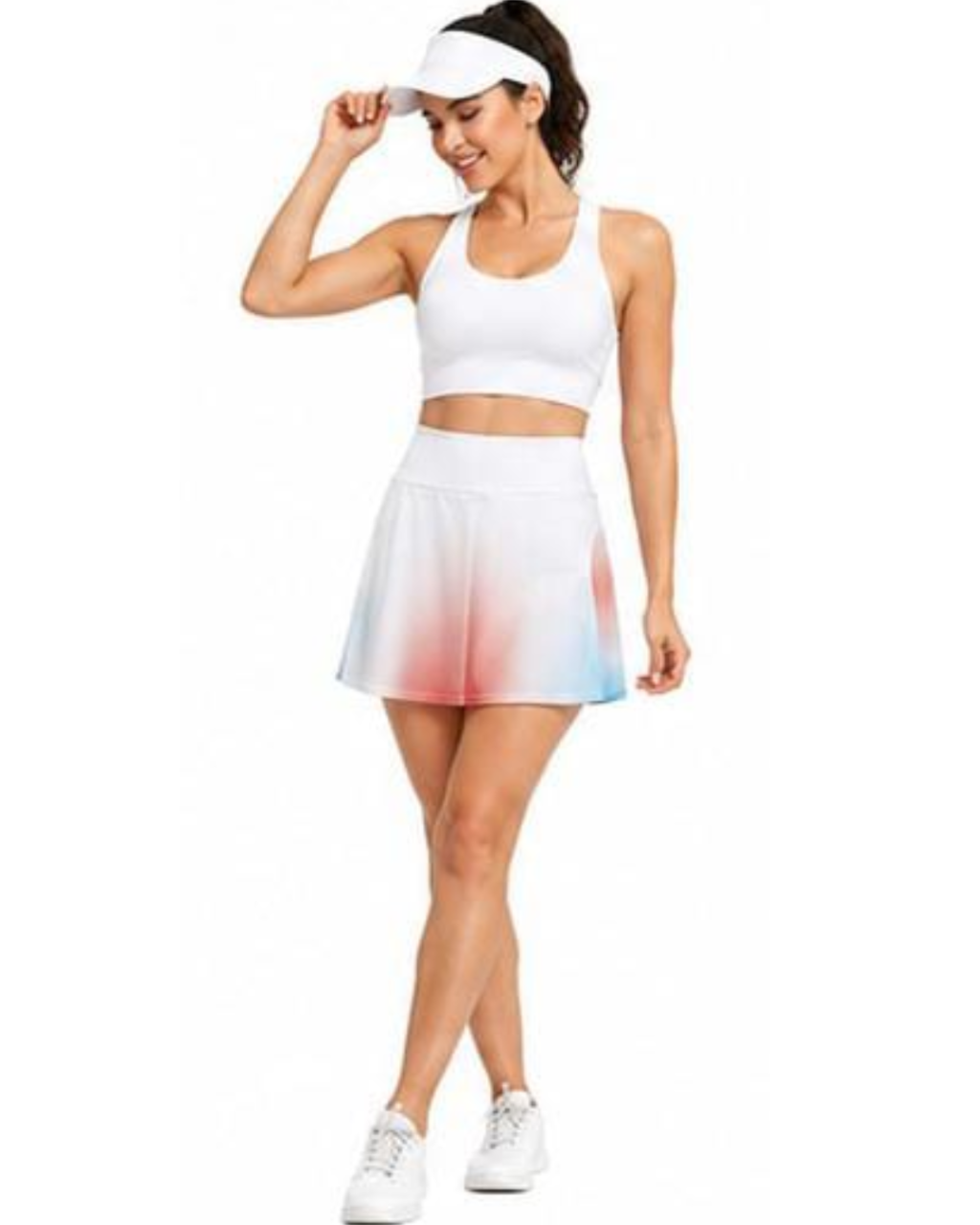 A woman modeling a white athletic set consisting of a sports bra, visor, and a white tennis skirt with a pink and blue ombre gradient at the hem, paired with white sneakers on a solid white background.