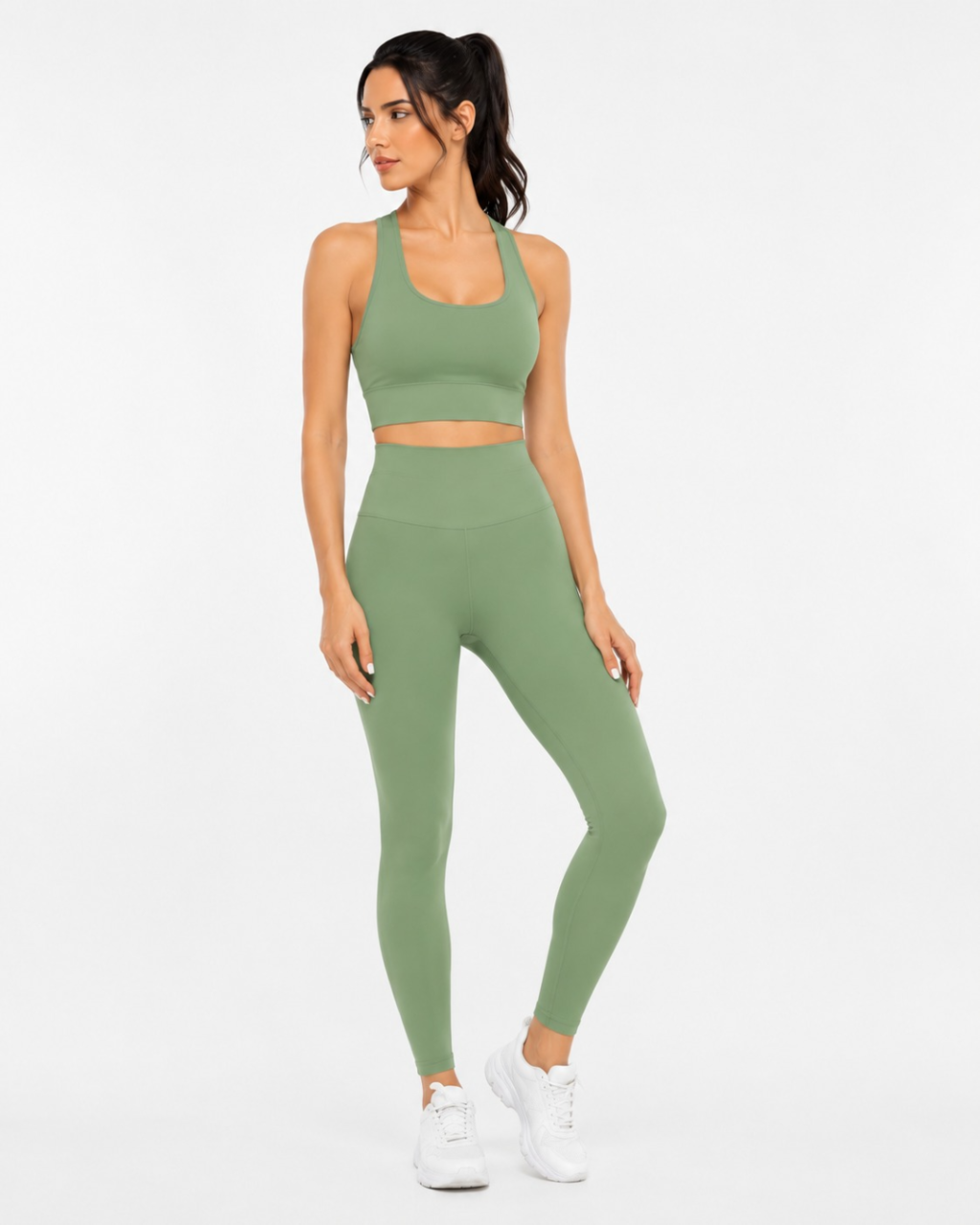 A woman modeling a matching sage green activewear set consisting of a sports bra and high-waisted leggings, paired with white sneakers, against a plain white studio background.