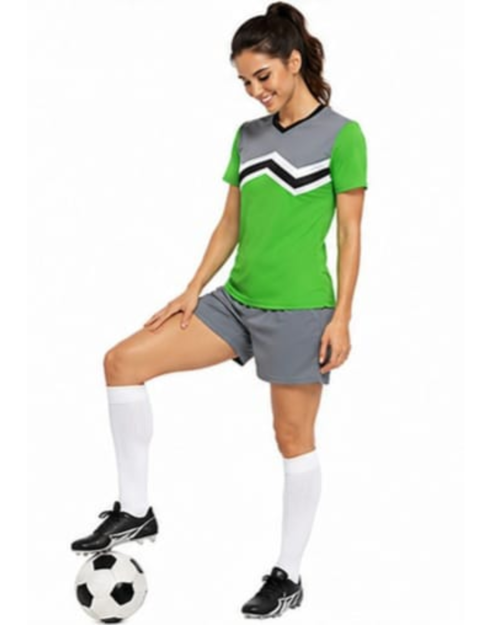 A woman modeling a green and gray soccer jersey with a black and white chevron pattern, gray shorts, white socks, and black cleats, standing on a soccer ball against a white background.