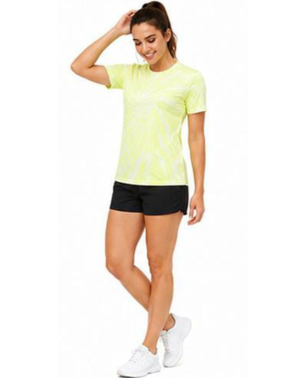A woman modeling a short-sleeve athletic t-shirt in neon yellow-green with a white tie-dye pattern, paired with black athletic shorts and white sneakers, against a plain white background.