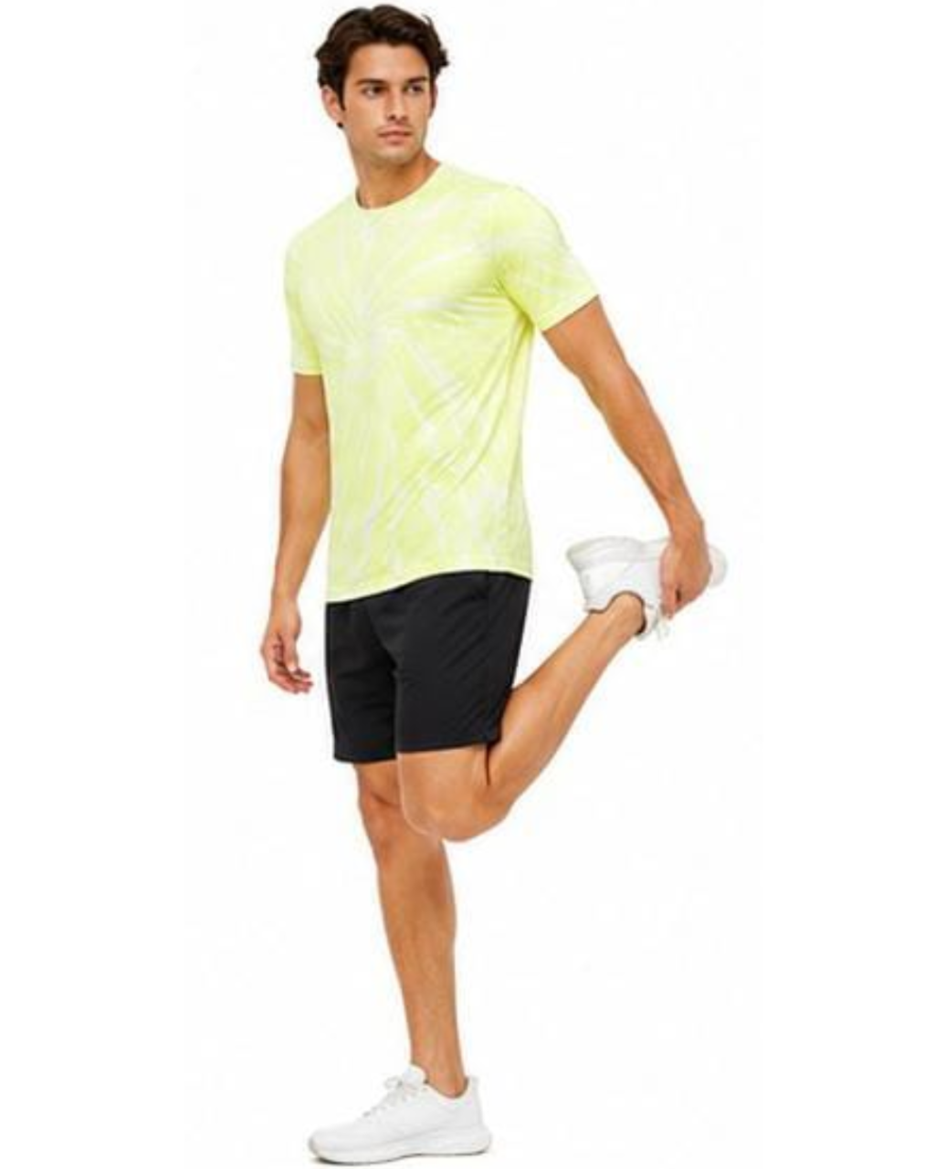 A man wearing a neon yellow-green tie-dye t-shirt, black athletic shorts, and white sneakers is stretching his leg against a plain white background.