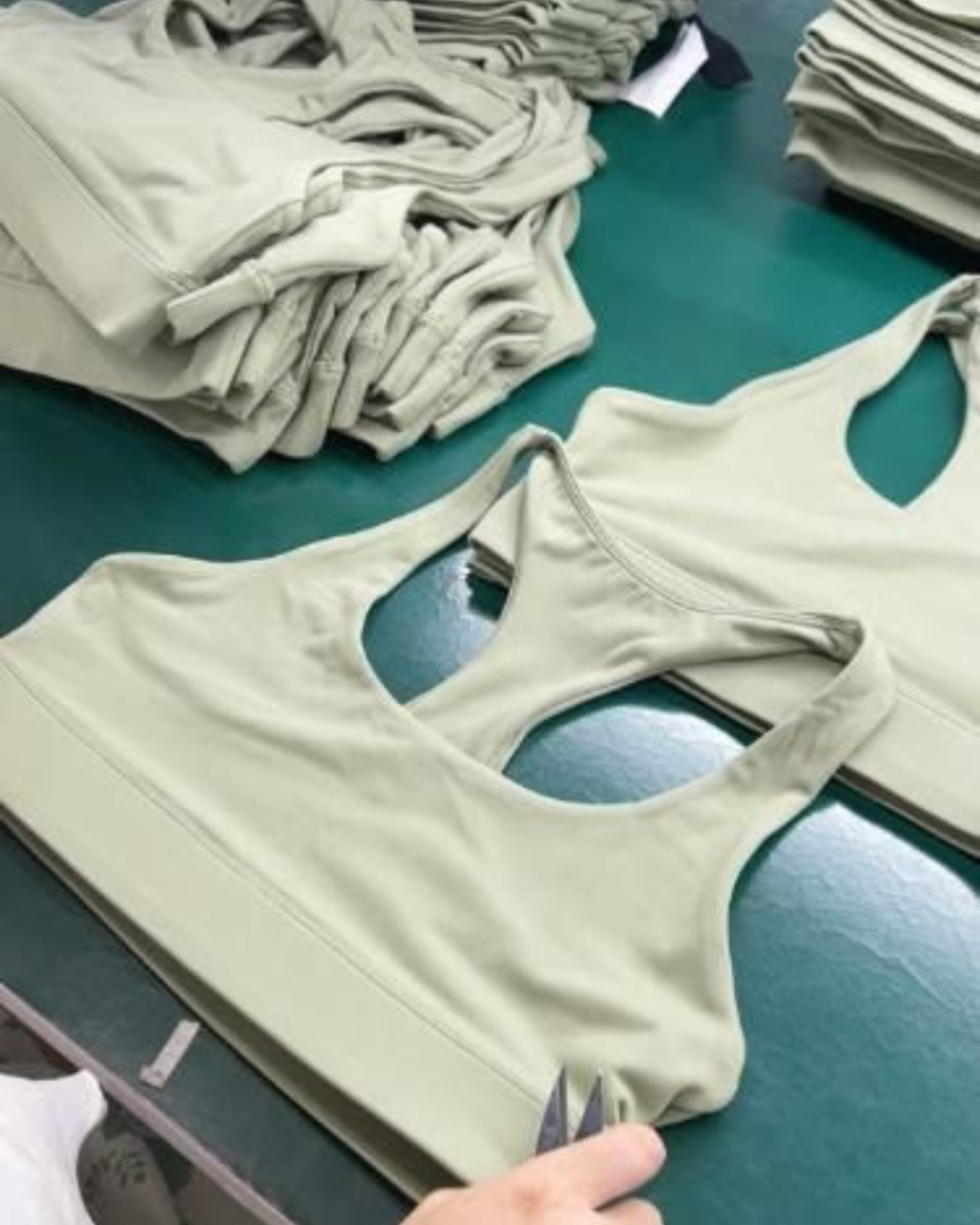 A sage green athletic sports bra laid flat on a green table, with folded matching tops stacked in the background. A hand is visible holding the edge of the garment.