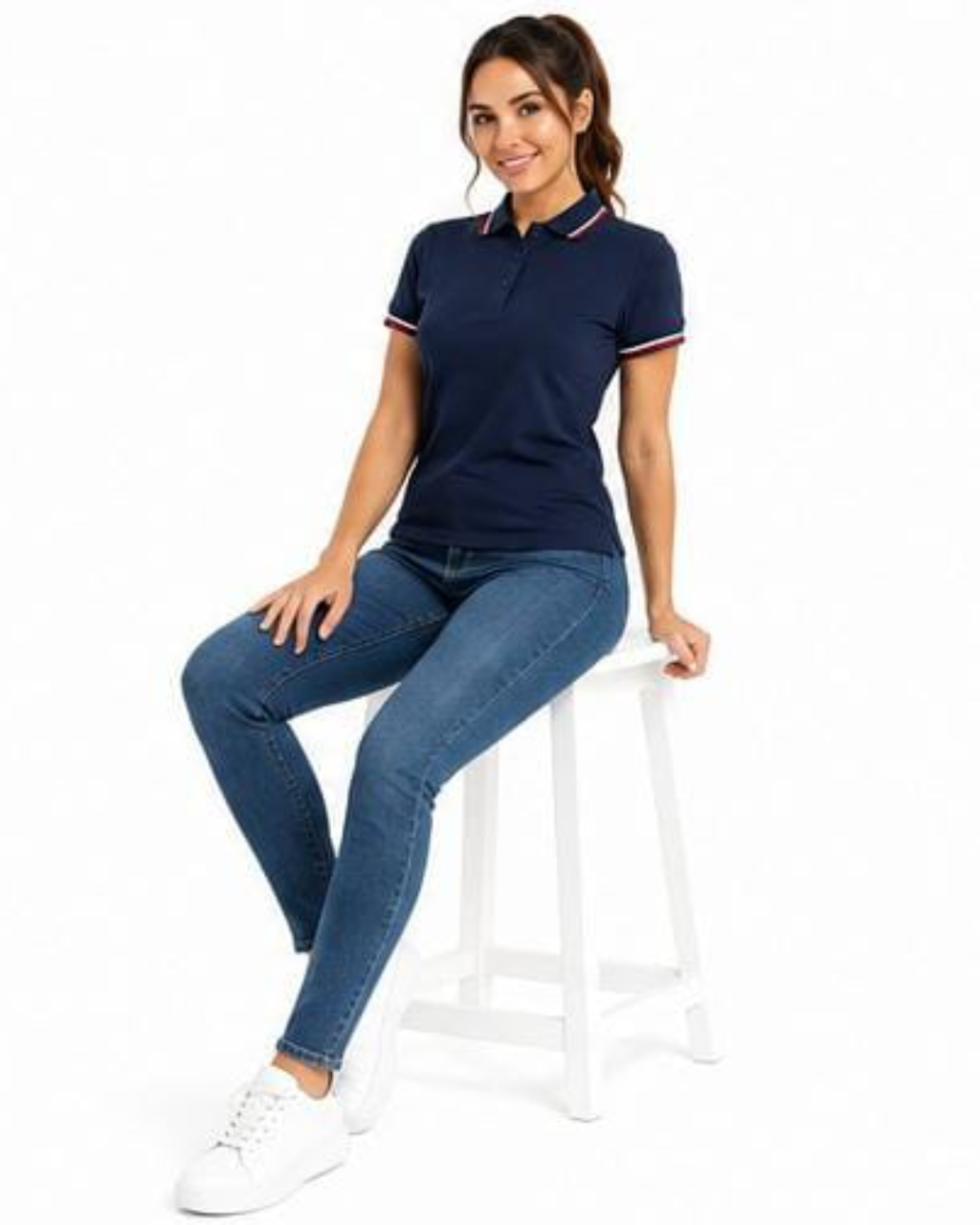 A woman wearing a navy blue short-sleeve polo shirt with white and red striped trim on the collar and sleeve cuffs, paired with blue jeans and white sneakers, sitting on a white stool against a white background.