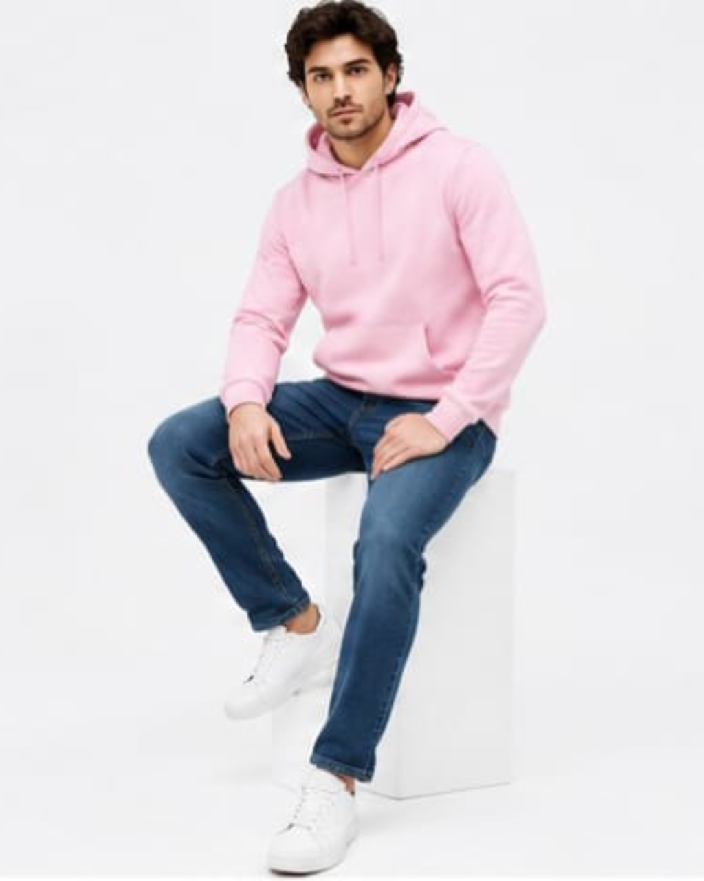 A man wearing a pink pullover hoodie, blue jeans, and white sneakers sits on a white block against a plain white studio background.