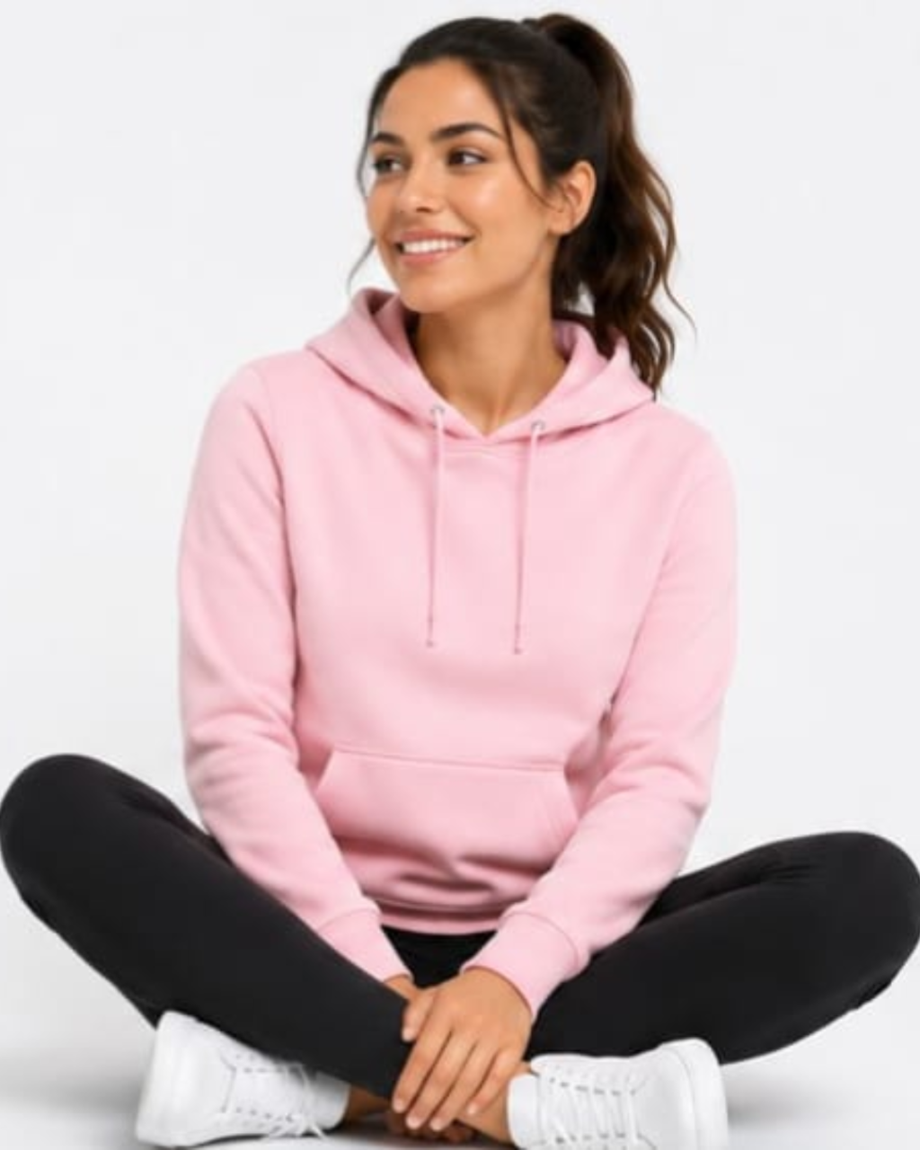 A woman sitting cross-legged wearing a light pink pullover hoodie, black leggings, and white sneakers against a plain white background.