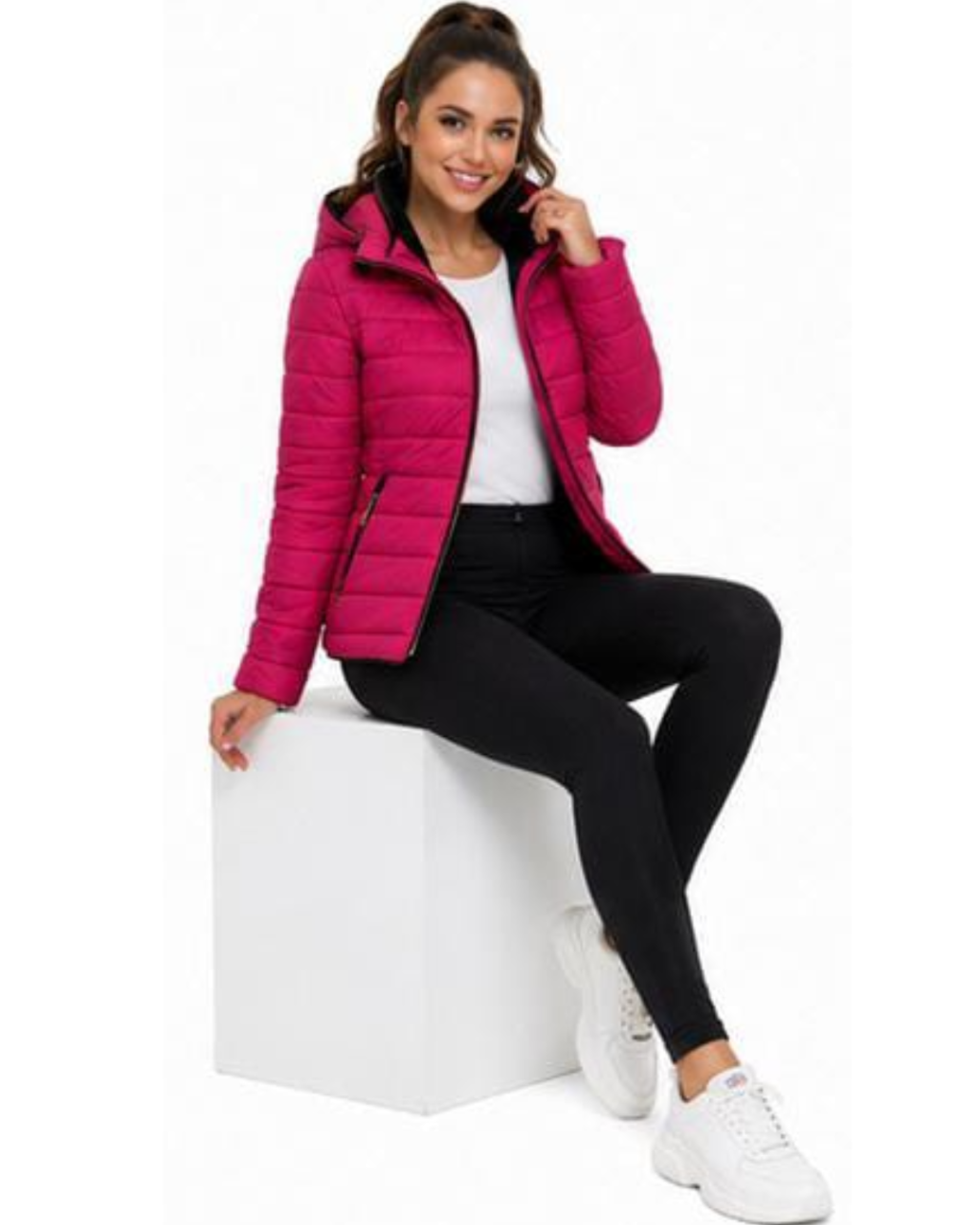 A woman wearing a bright pink puffer jacket, black leggings, and white sneakers sits on a white display block against a plain white background.