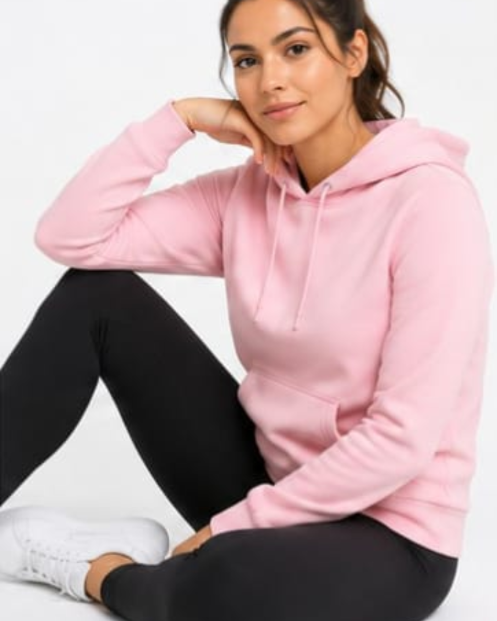 A woman wearing a light pink pullover hoodie and black leggings, sitting against a plain white background.