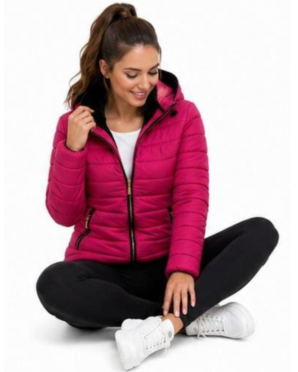 A woman sitting cross-legged on a white studio background wearing a bright pink puffer jacket, black leggings, and white sneakers.