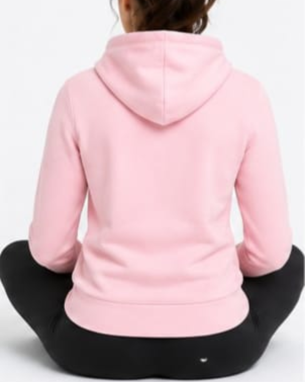A person sitting cross-legged facing away from the camera, wearing a light pink pullover hoodie and black leggings against a plain white background.