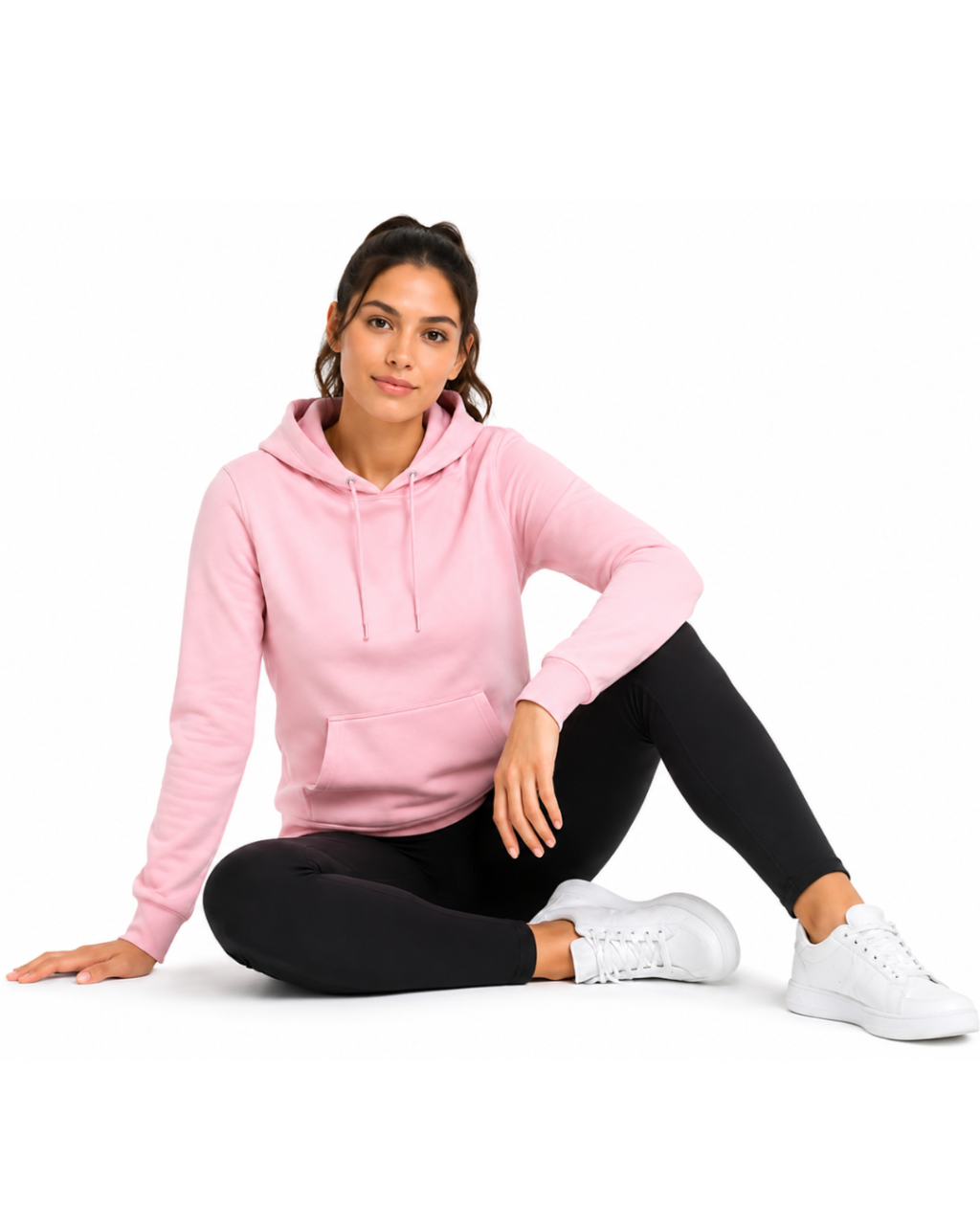 A woman sitting on a white studio floor wearing a light pink pullover hoodie, black leggings, and white sneakers.
