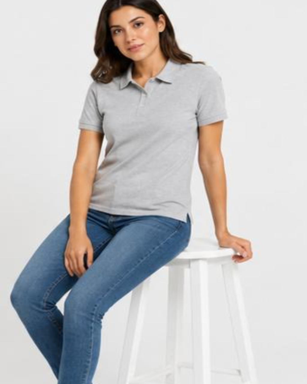A woman wearing a light gray short-sleeve polo shirt and blue jeans, sitting on a white stool against a light gray background.