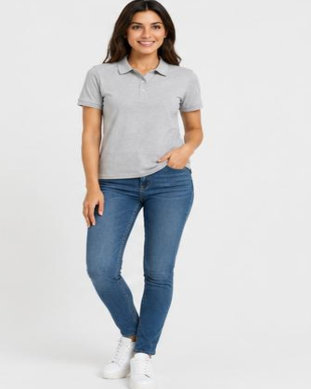 A woman modeling a light gray short-sleeve polo shirt with blue skinny jeans and white sneakers, standing against a plain white studio background.
