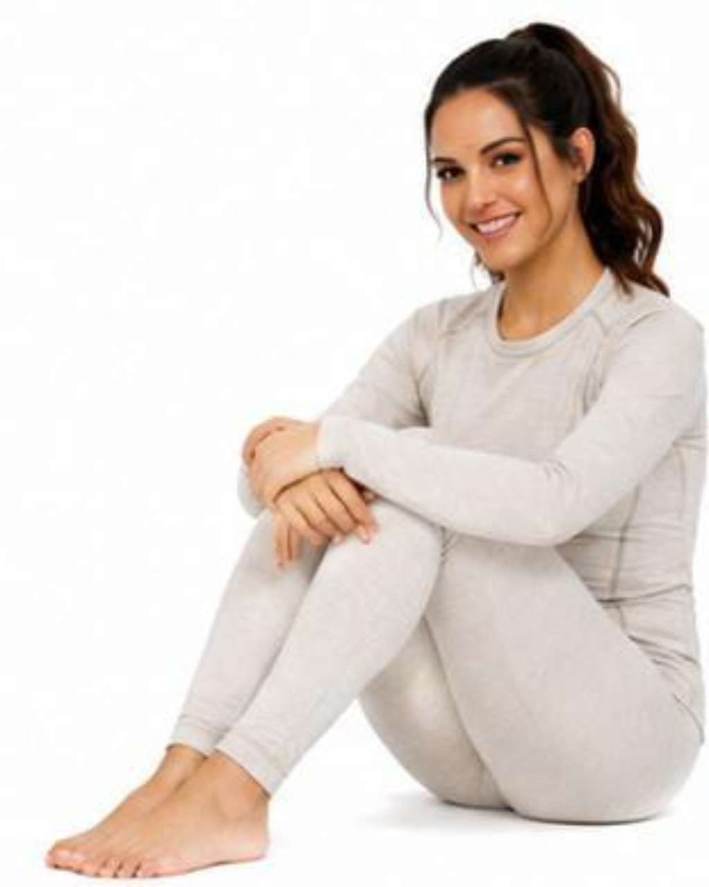 A woman sitting cross-legged on a white studio background wearing a light beige long-sleeve crewneck top and matching leggings.