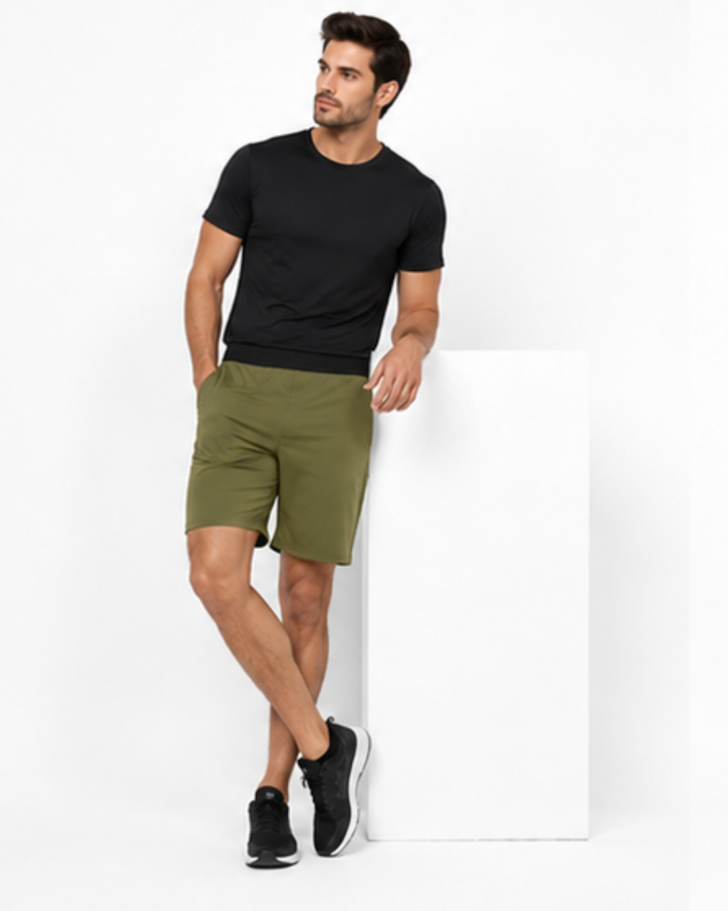 A man wearing a black short-sleeve t-shirt, olive green shorts, and black sneakers, leaning against a white display block in a studio setting.