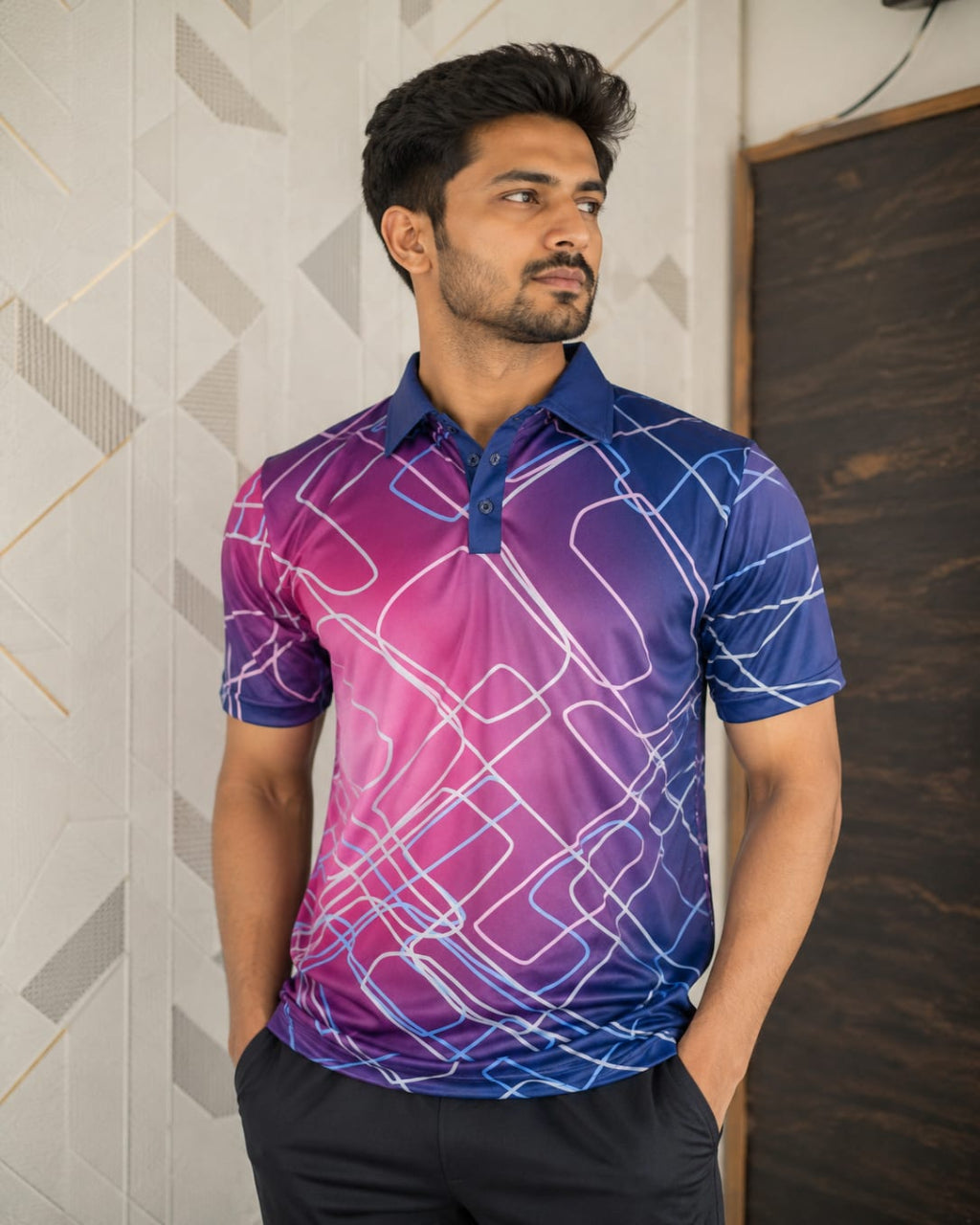 A man wearing a short-sleeve polo shirt with a blue-to-purple gradient and white geometric line pattern, standing indoors against a patterned wall.