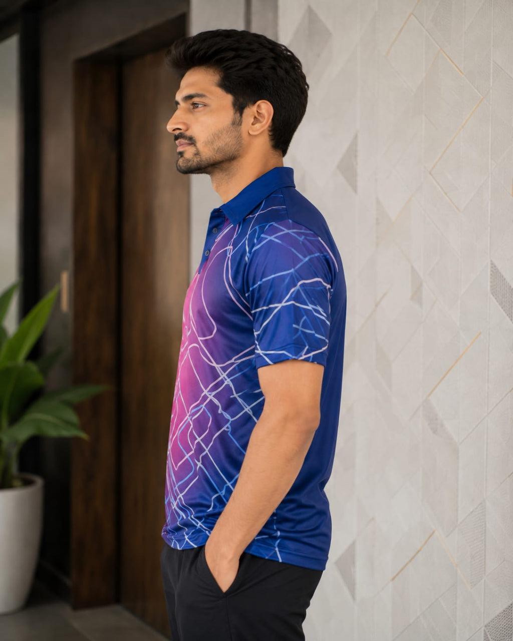 A man wearing a short-sleeve polo shirt with a blue-to-purple gradient and white abstract line pattern, standing in profile against a light textured wall.