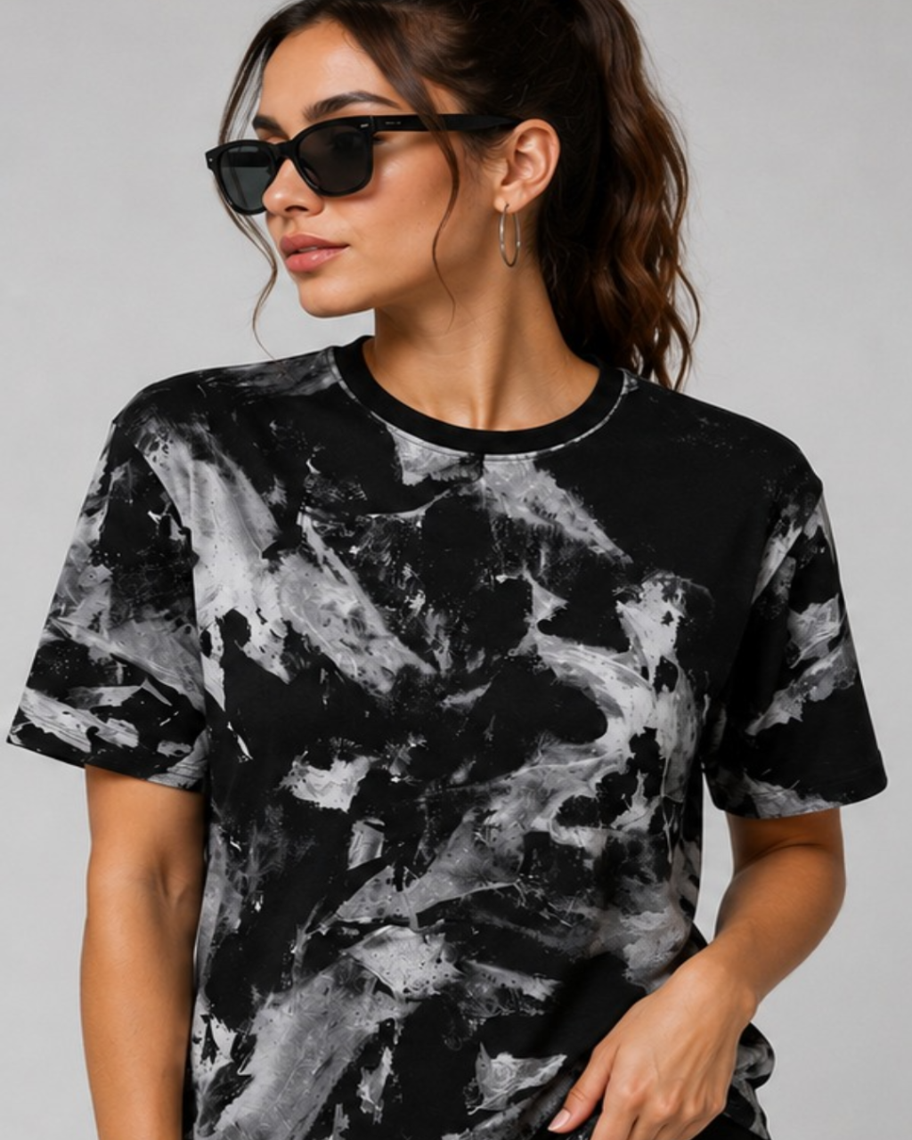 A woman wearing a black and gray abstract print t-shirt, black sunglasses, and hoop earrings, photographed against a solid light gray background.