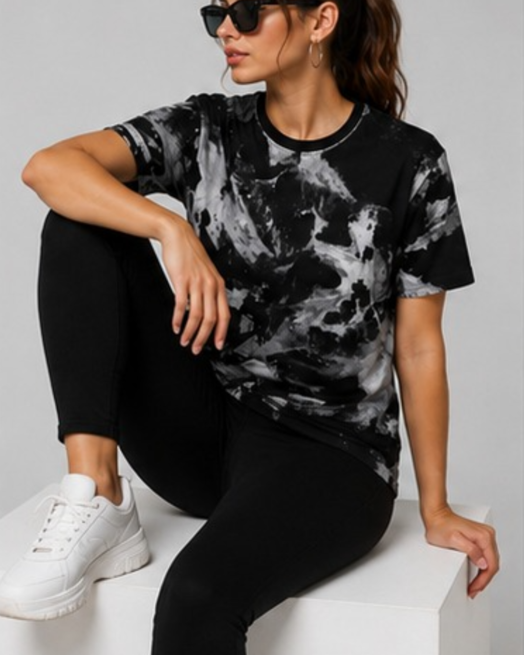 A woman wearing a black and gray marble-patterned t-shirt, black leggings, and white sneakers, sitting on a white block against a light gray background.