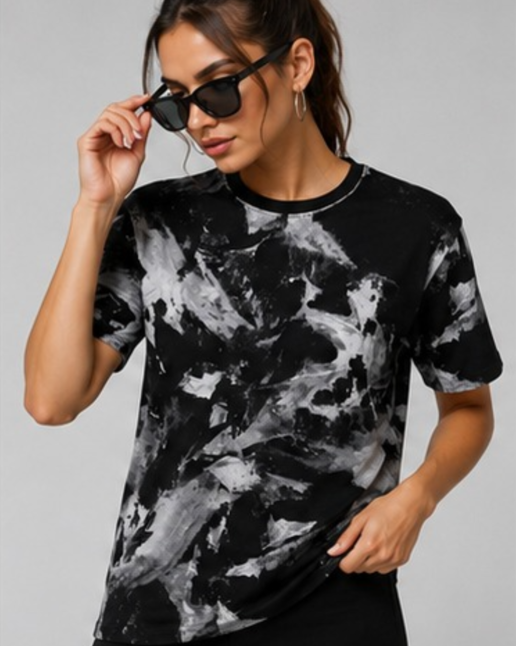 A woman wearing a black and white abstract marble print t-shirt and black sunglasses, posing against a solid gray studio background.