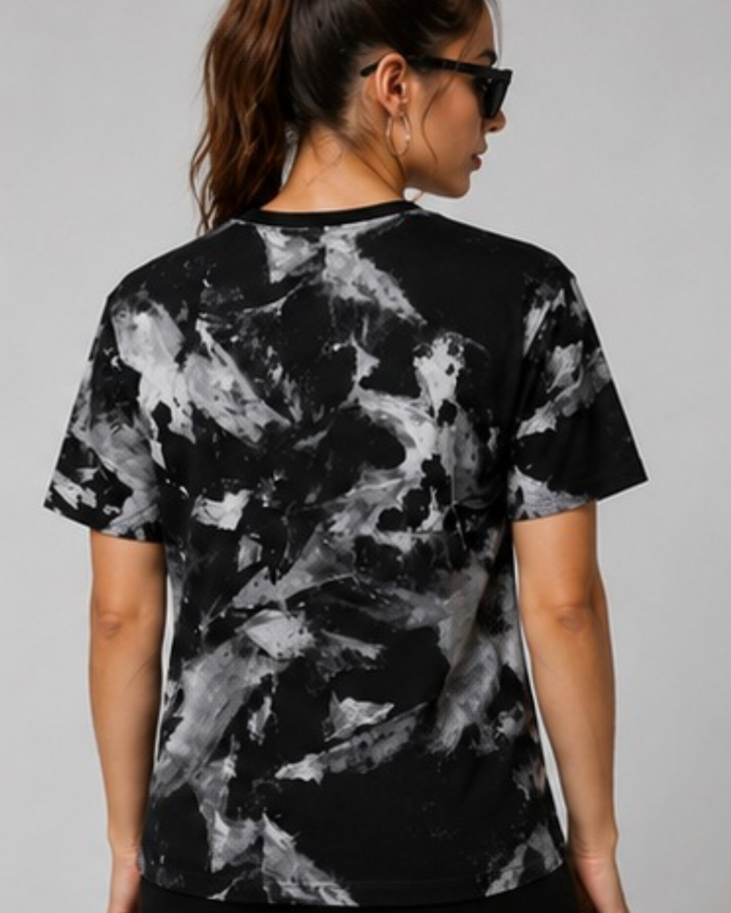 A woman shown from the back wearing a black short-sleeve t-shirt with a gray and white abstract marble or paint-splatter pattern, set against a plain light gray studio background.