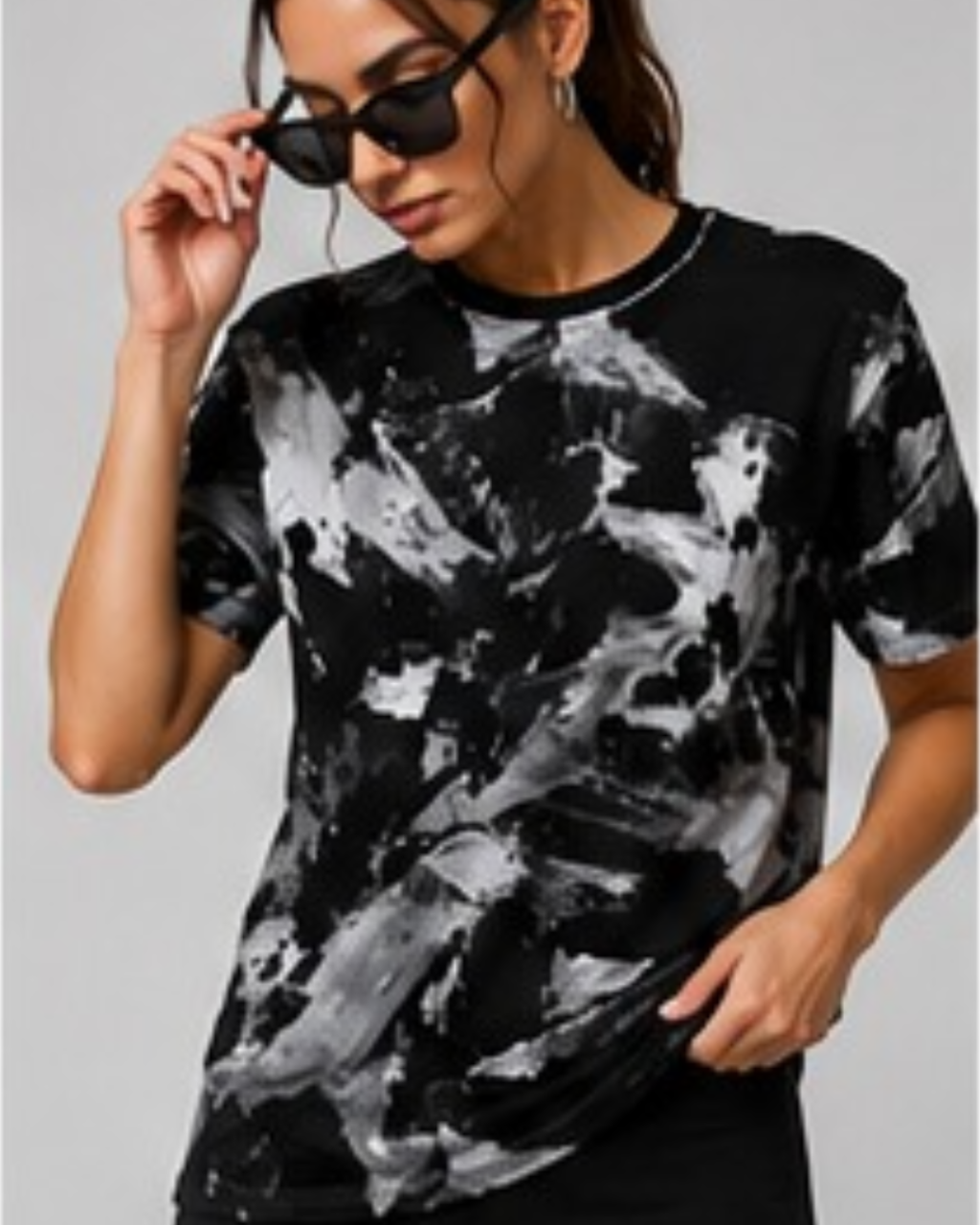 A woman wearing a black and white abstract tie-dye t-shirt and black sunglasses, posing against a solid gray background.