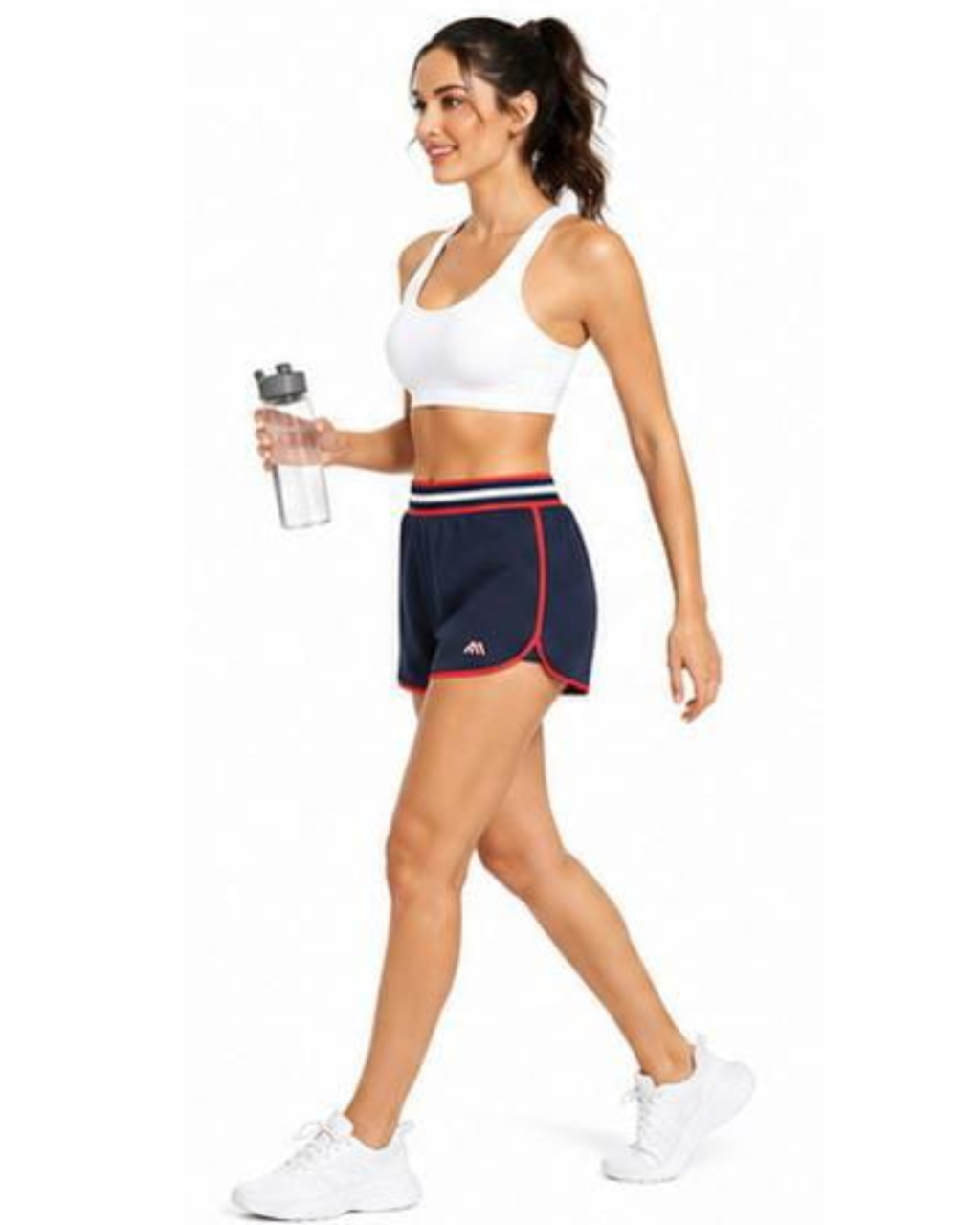 A woman modeling navy athletic shorts with red and white trim, a white sports bra, and white sneakers, holding a clear water bottle against a plain white background.