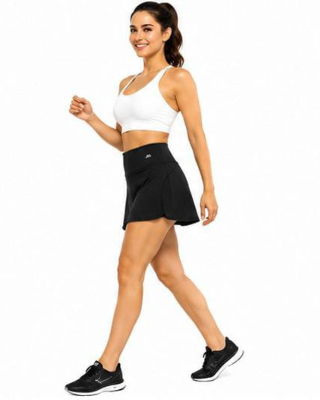 A woman modeling a white sports bra and black athletic skort with black running shoes, walking against a plain white background.