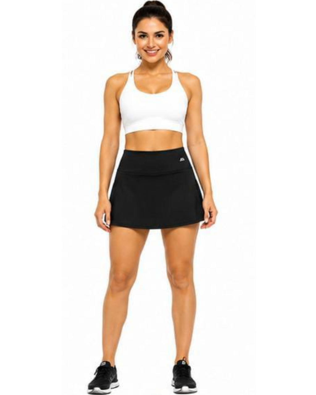 A woman modeling a white sports bra and a black athletic skort with a small white logo on the left hip, standing against a plain white background.