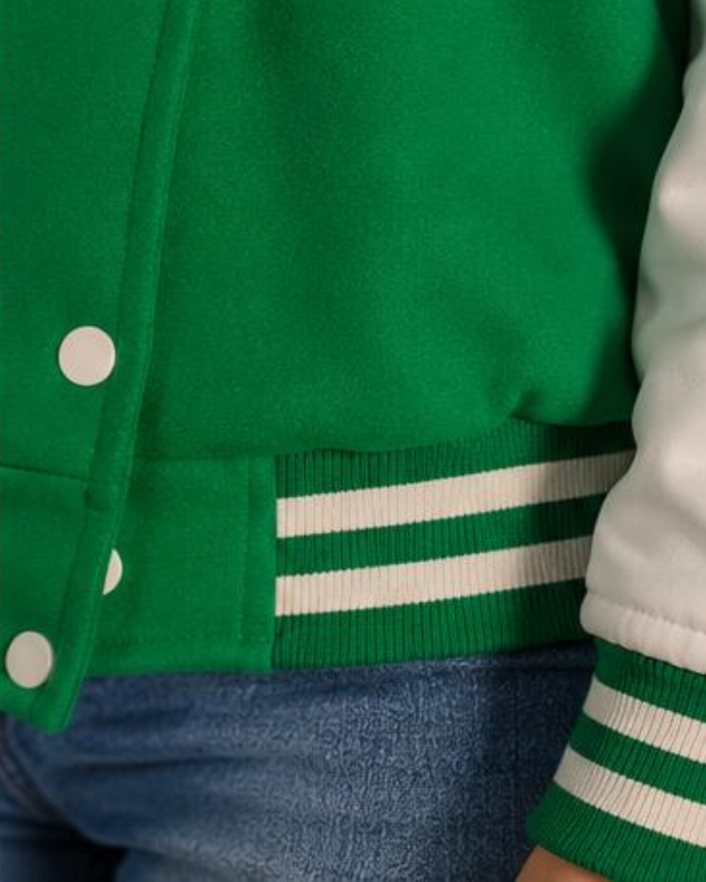 Green varsity jacket with white stripes on sleeve and buttons, worn over a white shirt and blue jeans.