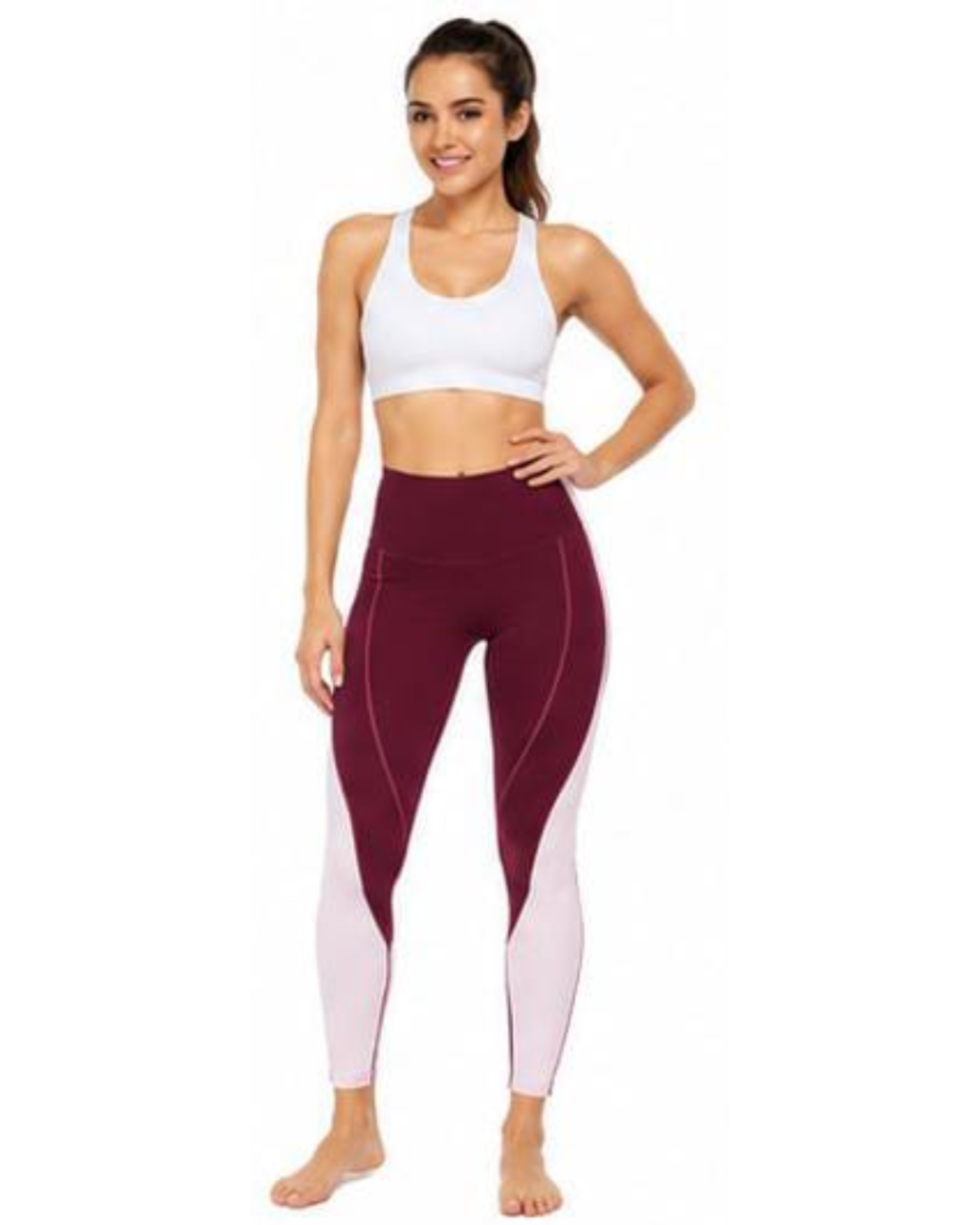 A woman modeling high-waisted colorblock leggings in burgundy and light pink, paired with a white sports bra, against a plain white background.