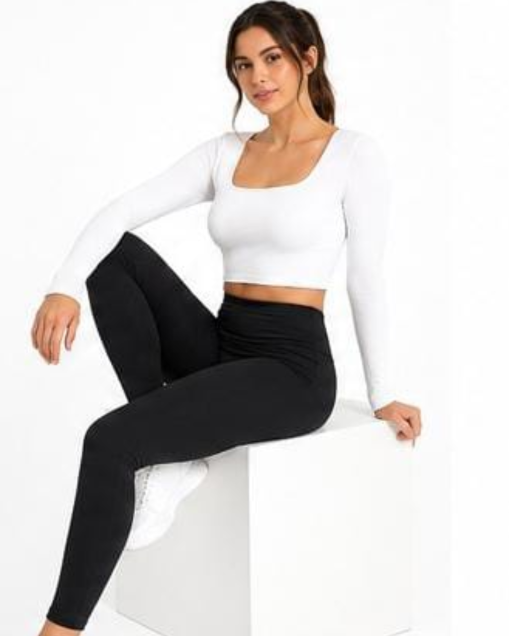 A woman sitting on a white block wearing a white long-sleeve crop top and black high-waisted leggings against a white background.