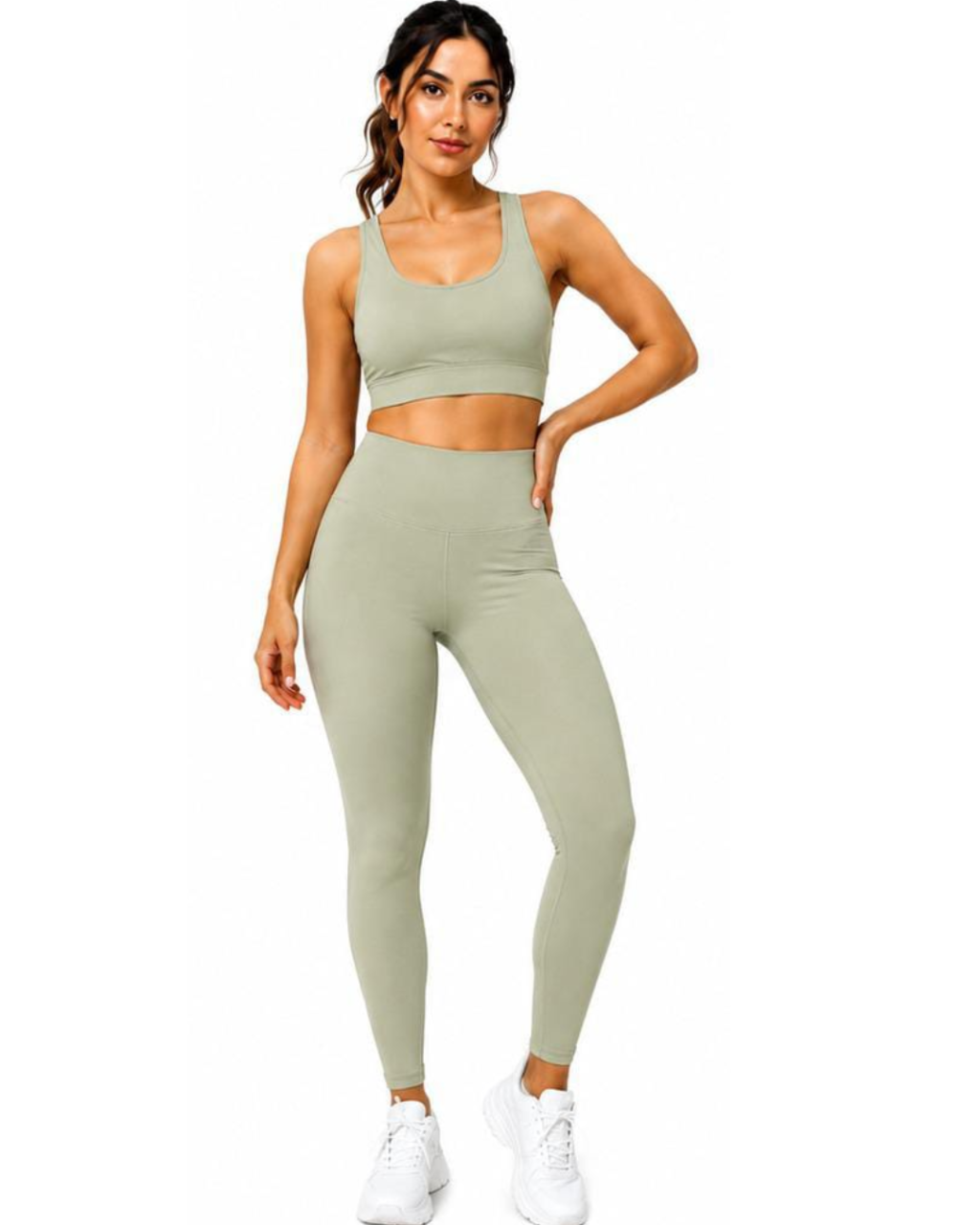 A woman modeling a matching sage green activewear set consisting of a sports bra and high-waisted leggings, paired with white sneakers, against a plain white background.