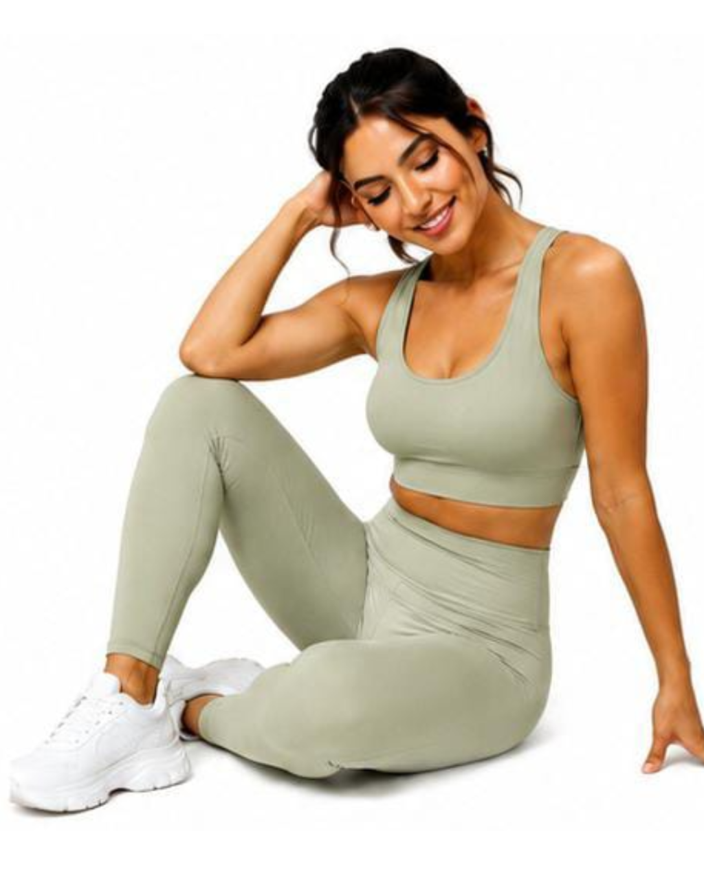 A woman modeling a matching sage green activewear set consisting of a sports bra and high-waisted leggings, paired with white sneakers, against a solid white background.