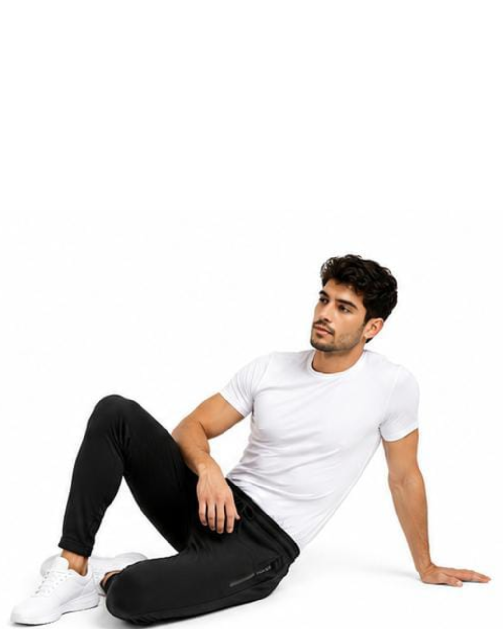A man wearing a plain white short-sleeve t-shirt, black jogger pants, and white sneakers, sitting on a white studio floor against a white background.
