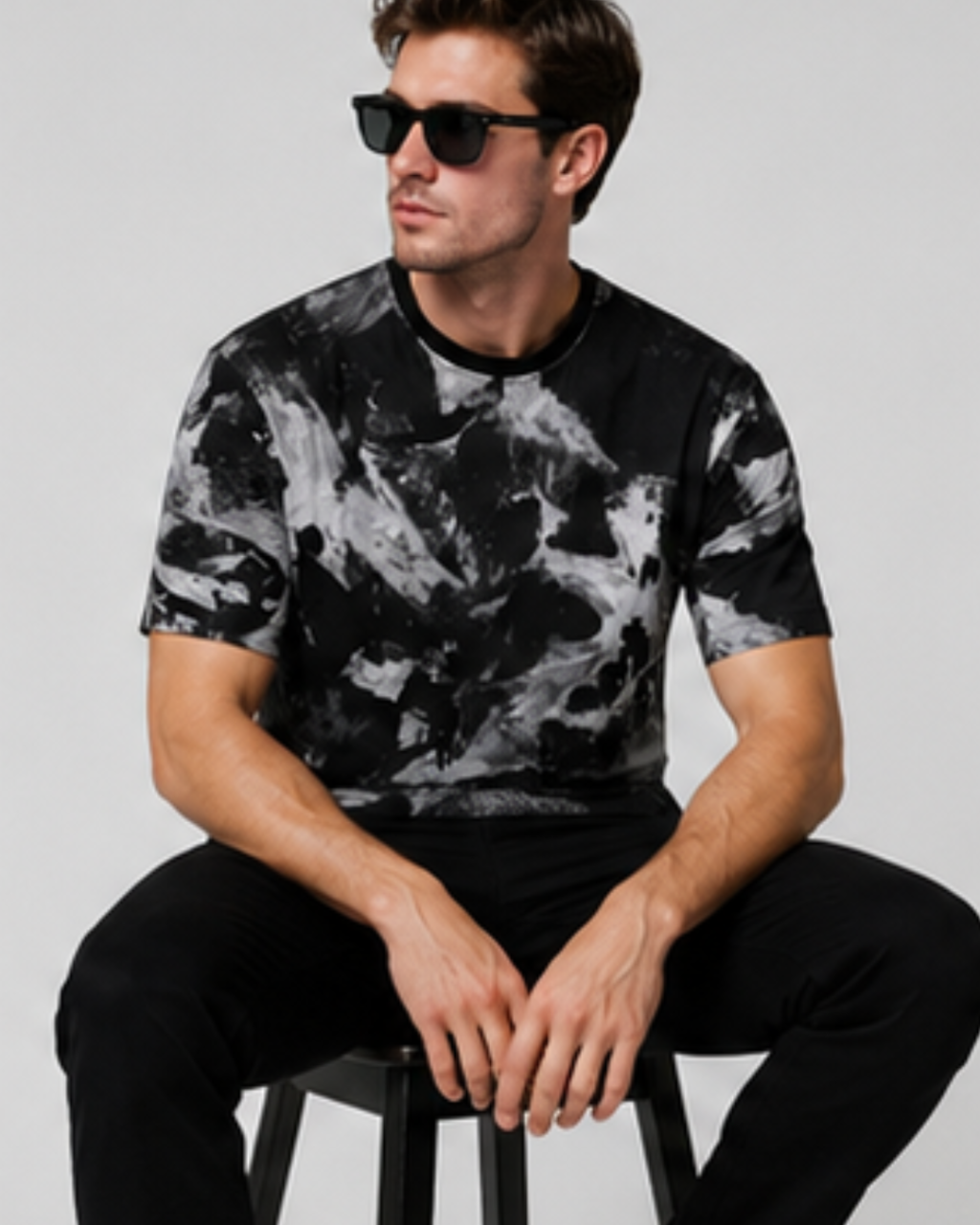 A man wearing a black and white abstract print t-shirt and black pants, sitting on a stool against a light gray background.