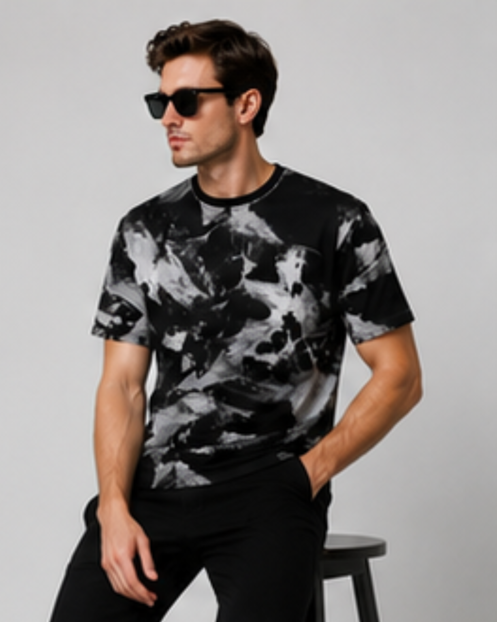 A man wearing a black and gray abstract print t-shirt, black pants, and black sunglasses, standing next to a stool against a light gray background.