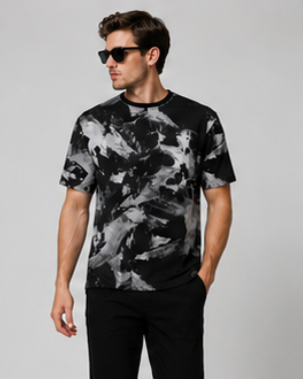 A man wearing a black and gray abstract painterly print t-shirt, black pants, and black sunglasses, standing against a light gray studio background.