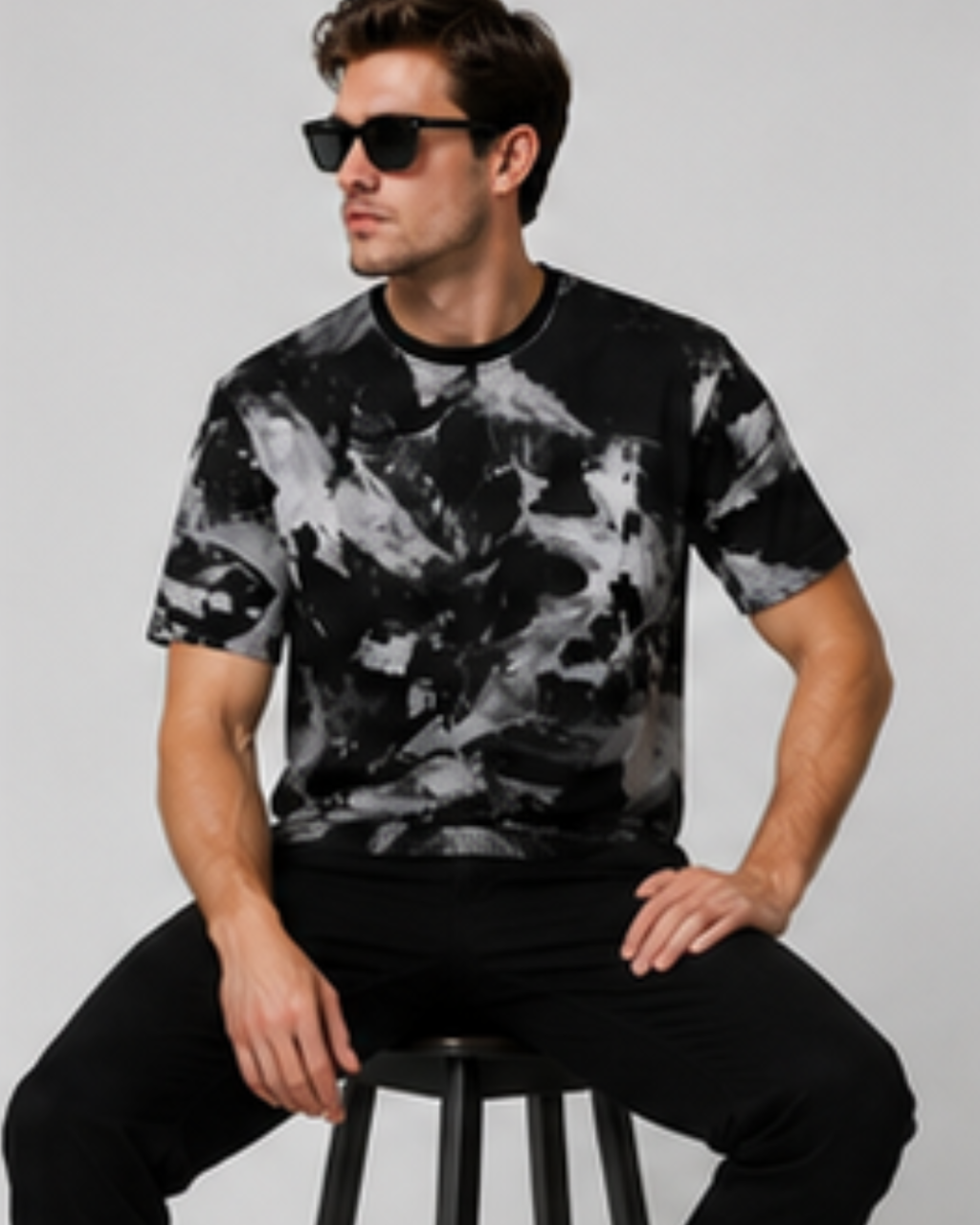 A man wearing a black and white abstract patterned t-shirt and black pants, sitting on a stool against a light gray background.