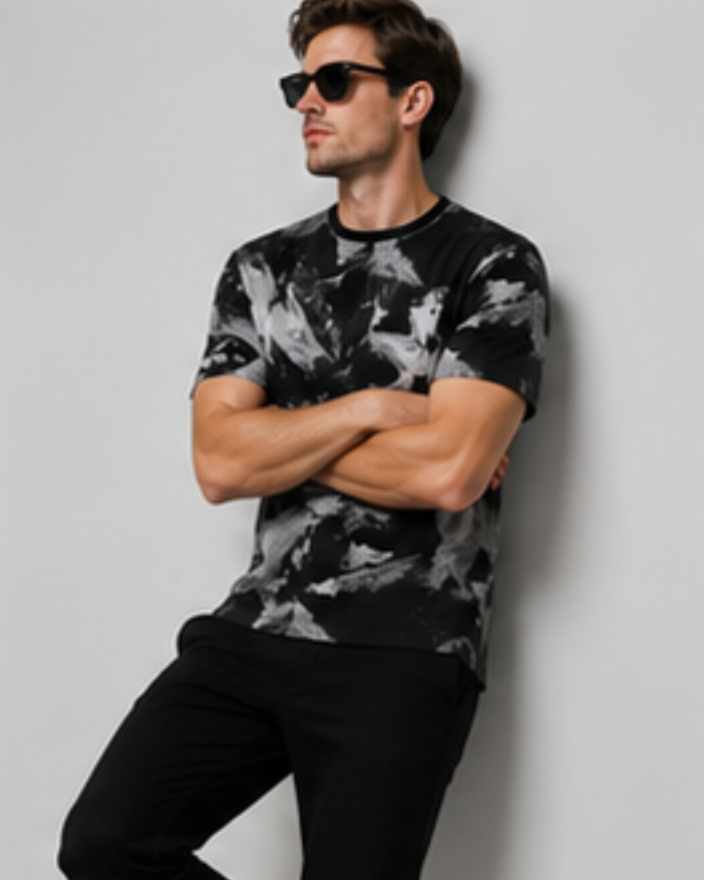 A man wearing a black and gray abstract patterned t-shirt, black pants, and black sunglasses, posing against a light gray wall.