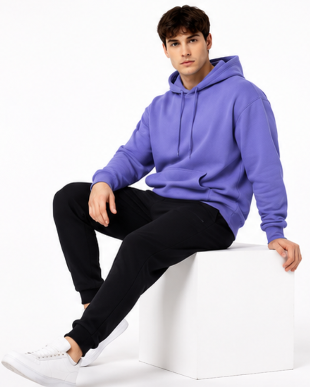 A man wearing a purple pullover hoodie, black sweatpants, and white sneakers sits on a white cube against a plain white studio background.