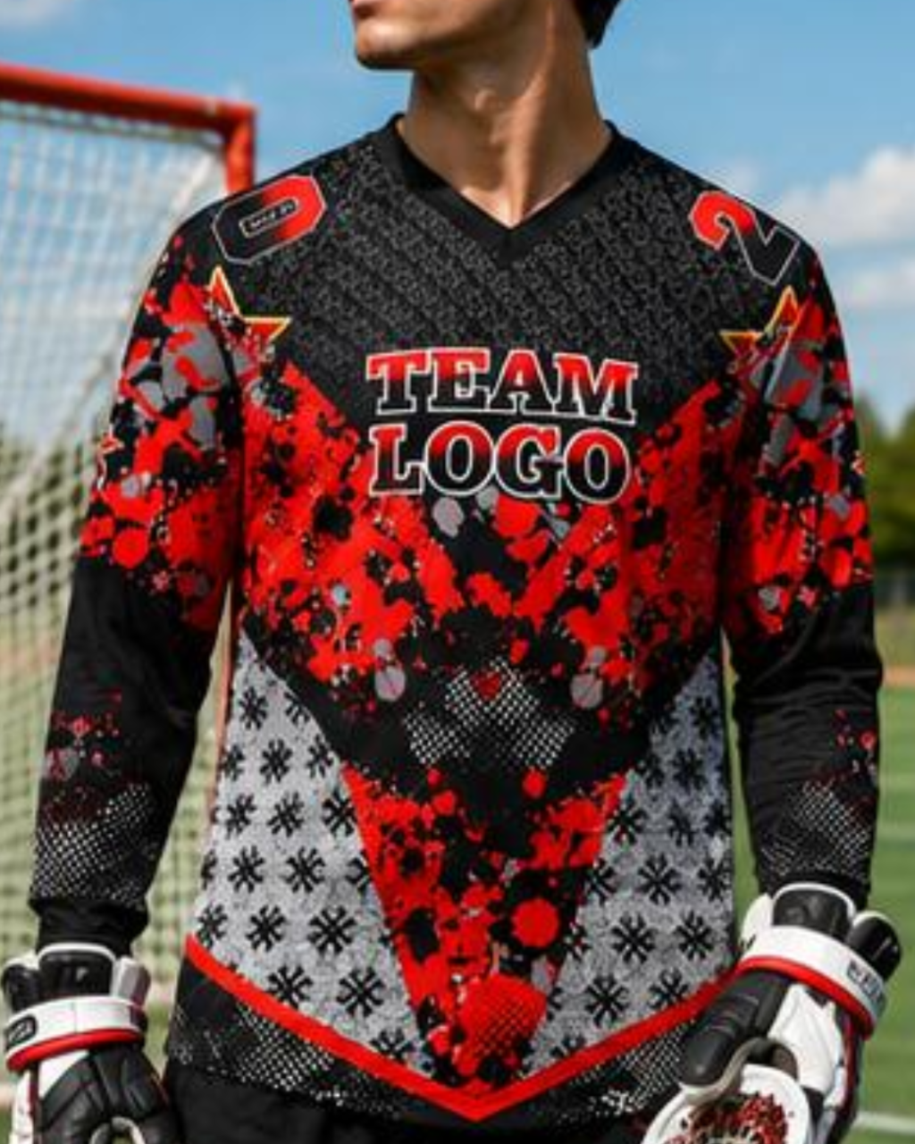 A man wearing a long-sleeve soccer goalkeeper jersey with a red, black, and gray patterned design, featuring 'TEAM LOGO' printed on the chest. He is also wearing white and black goalie gloves, with a soccer field and goal net visible in the background.