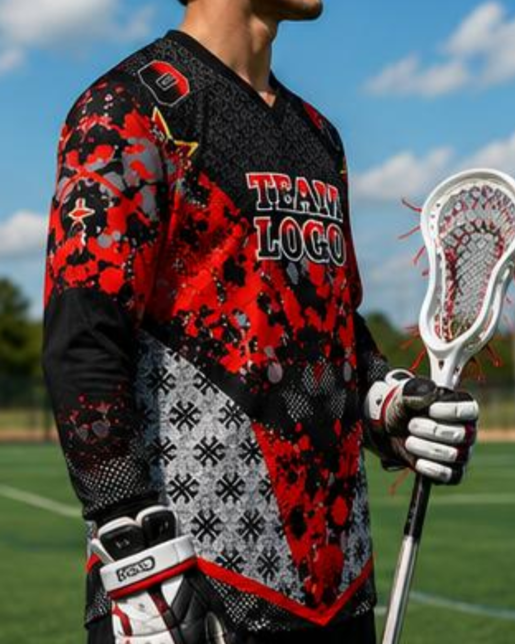 A close-up of a person wearing a long-sleeve lacrosse jersey outdoors on a field. The jersey is black and red with a splatter pattern, gray snowflake motifs on the lower front, and 'TEAM LOGO' printed across the chest.
