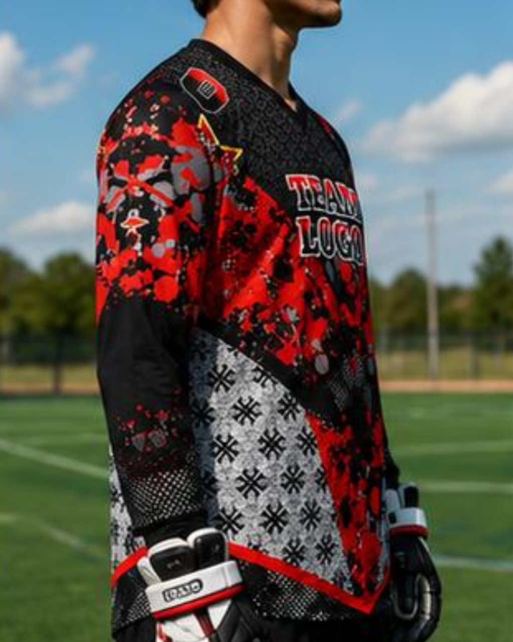A close-up side view of a person wearing a long-sleeve athletic jersey on an outdoor sports field. The jersey has a bold black, red, and gray pattern with the text 'TEAM LUCAS' on the chest and a visible 'GRAND' logo on the glove.