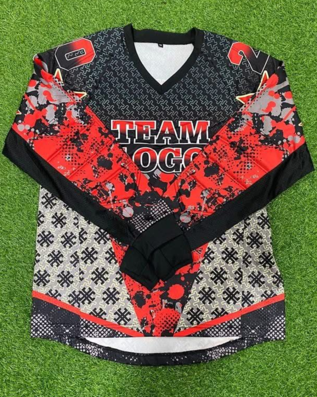 A long-sleeve hockey-style jersey laid flat on artificial green turf. The jersey has a black and red camouflage pattern with a black-and-white geometric pattern on the lower section, a black V-neck collar, and 'TEAM LOGO' printed across the chest.
