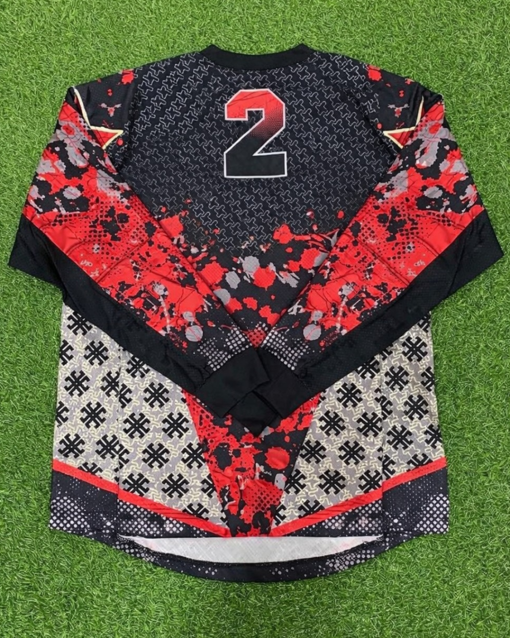 A long-sleeve hockey jersey laid flat on green artificial turf, featuring a black and red abstract pattern with a large number 2 on the back.