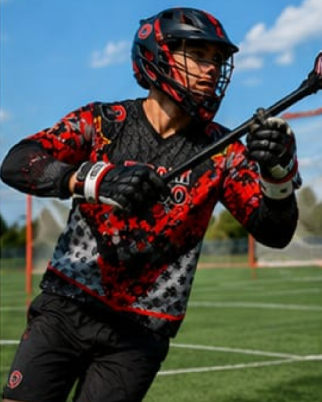 A lacrosse player wearing a red, black, and gray patterned jersey, black helmet, and gloves on an outdoor sports field under a blue sky.